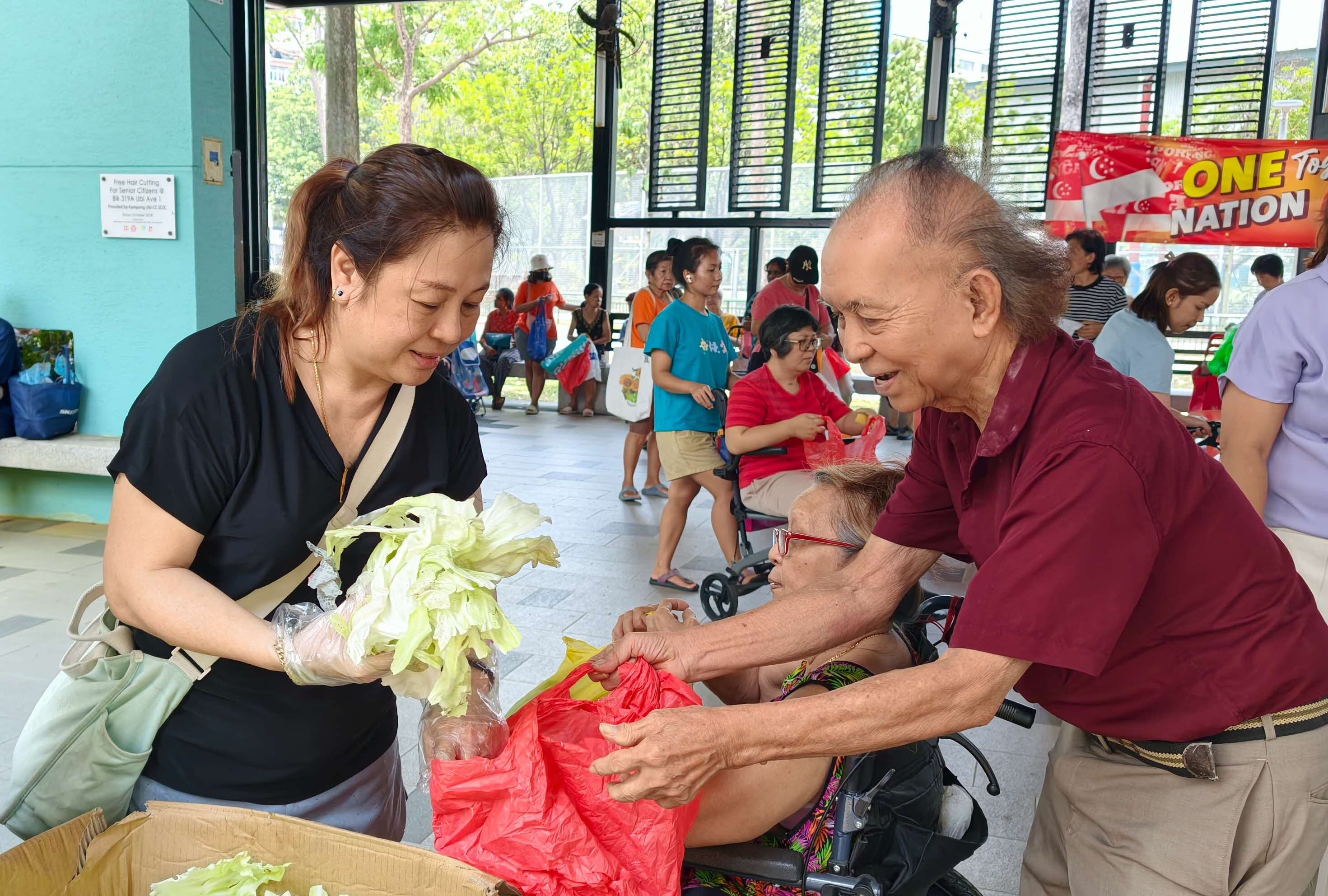 A volunteer distributes vegetables to an elderly man and a wheelchair-user senior at a community distribution event.
